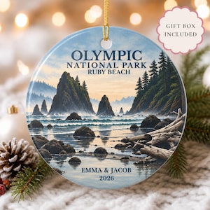 May include: A round ceramic ornament featuring a landscape illustration of Olympic National Park Ruby Beach with sea stacks, trees, and driftwood. The text reads OLYMPIC NATIONAL PARK RUBY BEACH, EMMA & JACOB, and 2026. A gold string is attached for hanging.