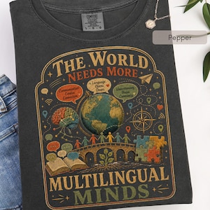 May include: A charcoal gray t-shirt featuring a graphic print with the text "THE WORLD NEEDS MORE MULTILINGUAL MINDS". The design includes a globe, a brain, a bridge with people, an open book, puzzle pieces, and speech bubbles with text.