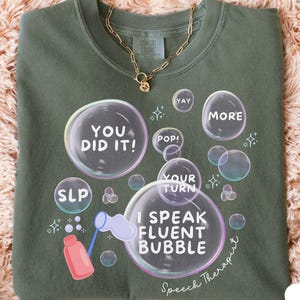May include: Moss green Comfort Colors t-shirt with a bubble-themed graphic. The design includes text bubbles with phrases like "YOU DID IT!" and "I SPEAK FLUENT BUBBLE." A gold chain necklace with a pendant is draped on the shirt.