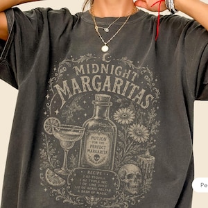 May include: A charcoal gray t-shirt featuring a graphic print with the text "MIDNIGHT MARGARITAS" and "POTION FOR THE PERFECT MARGARITA". The design includes a margarita glass, a bottle, a skull, flowers, and a recipe listing 2 oz tequila.