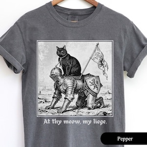 May include: Dark grey t-shirt featuring a black and white illustration of a black cat riding a knight in armor. The knight is on his hands and knees, holding a flag. The text "At thy meow, my liege" is below the image.