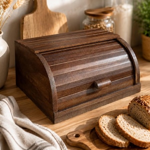 Puede incluir: Una panera de madera de color marrón oscuro con una tapa enrollable de listones horizontales y un pequeño tirador rectangular. La panera tiene una base rectangular y una parte superior curva, fabricada en madera teñida con veta visible.