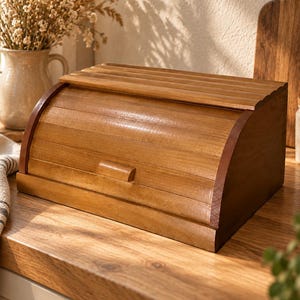 May include: A medium-brown wooden bread box featuring a curved, slatted roll-top lid with a small rectangular handle. The box has a smooth, polished finish and a rectangular base, designed to store baked goods on a kitchen countertop.