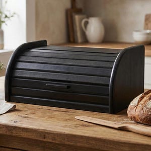 May include: A black wooden bread box with a roll-top lid and a small handle. The box is on a wooden countertop next to a loaf of bread and a wooden knife. The kitchen setting includes a bowl of fruit and a window.