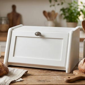 Puede incluir: Una panera de madera blanca con la parte superior curvada y un pequeño tirador metálico redondo. La panera está sobre una mesa de madera junto a una hogaza de pan y una tabla de cortar. La panera mide unos 38 cm de ancho.