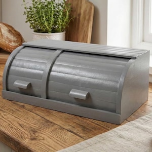 May include: A gray wooden bread box with a roll-top design and two small handles. The bread box is on a wooden countertop, with a loaf of bread and a butter dish in the background. The bread box is a kitchen storage item.