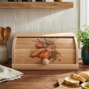 May include: A wooden bread box with a roll-top lid. The front features a painted illustration of bread loaves and lavender. The box has a wooden knob and is set on a wooden surface. Kitchen storage.