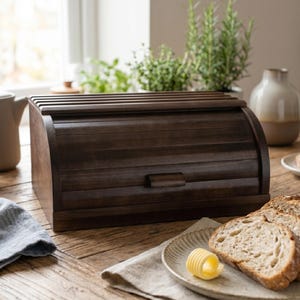 May include: A dark brown wooden bread box with a roll-top lid. The box is rectangular with a curved front and a small handle. Sliced bread and a pat of butter are on a plate in front of the box. The scene is set on a wooden table.