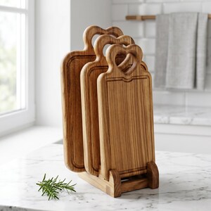 May include: A set of three wooden cutting boards with heart-shaped handles, displayed in a wooden stand. The boards have a rectangular shape with a groove around the edge. The wood has a warm, natural tone, and the stand is made of the same material.