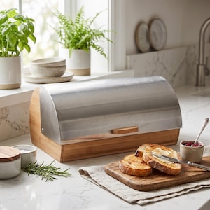 Könnte beinhalten: Ein Brotkasten mit einem hellbraunen Bambussockel und einem geschwungenen Roll-Top-Deckel aus Edelstahl mit kleinem Holzgriff. Zwei Scheiben geröstetes Brot liegen auf einem Holzschneidebrett neben einem Buttermesser und einer kleinen Schale.