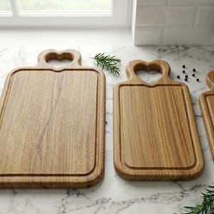 May include: Three wooden cutting boards with heart-shaped handles. The boards are rectangular with a groove around the edge. The wood grain is visible, and the boards are light brown. Rosemary sprigs and peppercorns are scattered around the boards.