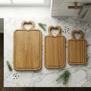 May include: Three wooden cutting boards with heart-shaped handles. The boards are different sizes and have a rectangular shape with a groove around the edge. The wood grain is visible, and the boards are arranged on a marble countertop.