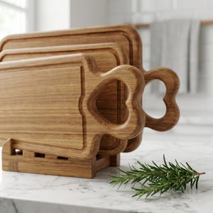 May include: A set of three wooden cutting boards with heart-shaped handles. The boards are made of light brown wood and are stacked in a wooden holder. A sprig of rosemary rests on the counter in front of the boards.
