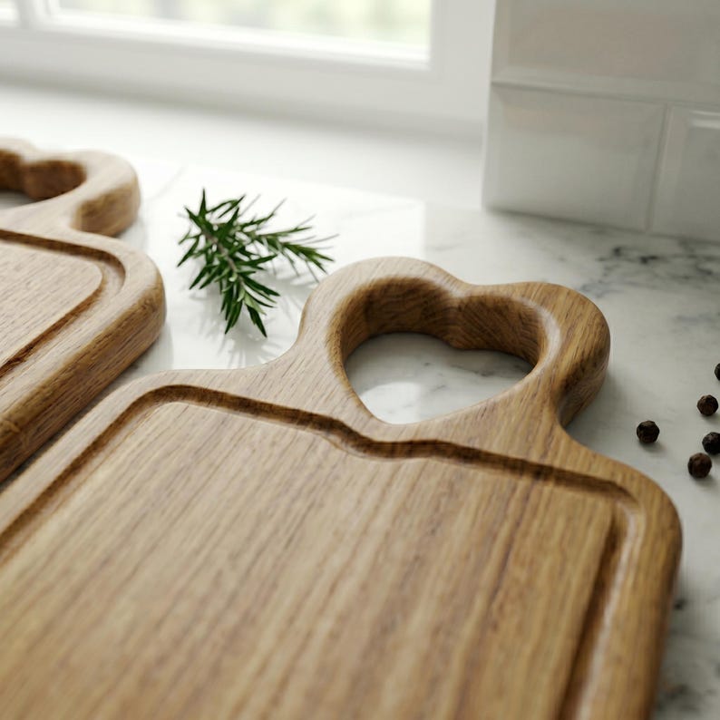 May include: Two wooden cutting boards with heart-shaped handles. The boards have a rectangular shape with a recessed area for food. A sprig of rosemary and peppercorns are also visible.