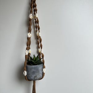 Op de afbeelding: Een bruine macramé plantenhouder met witte en gele bloemaccenten, met een grijze metalen pot met een groene vetplant. De houder heeft een houten ring aan de bovenkant en een kwast aan de onderkant.