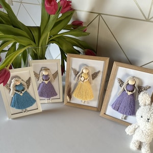 Op de afbeelding: Vier ingelijste engelenkunstwerken met houten frames. Elke engel heeft een houten hoofd, garen haar en een garen jurk in blauw, paars en geel. De engelen houden harten vast en hebben houten vleugels. Een boeket roze tulpen staat op de achtergrond.