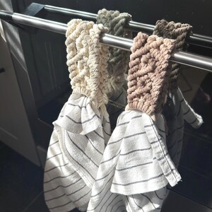 Op de afbeelding: Drie keukendoeken hangen aan een roestvrijstalen ovenstang. De handdoeken hebben een wit en zwart gestreept patroon. De handdoekhouders zijn gemaakt van geweven beige, groen en bruin touw.