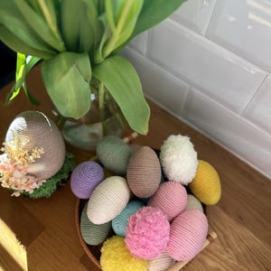 Op de afbeelding: Een verzameling kleurrijke, decoratieve paaseieren in een gevlochten mand. De eieren zijn in verschillende kleuren, waaronder roze, geel, groen en paars. Een groot, decoratief ei ligt op mos. Een vaas met tulpen staat op de achtergrond.