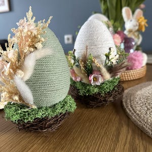 Op de afbeelding: Twee decoratieve paasei-ornamenten. Eén is lichtgroen met gedroogde bloemen en de andere is wit met bloemaccenten. Beide eieren zijn geplaatst in kleine, bruine takkennesten met groen mos.