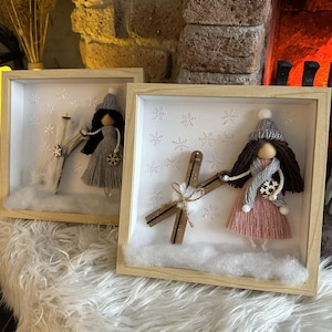 Könnte beinhalten: Zwei gerahmte Winter-Schattenboxen. Jede zeigt eine Puppe mit einem Kleid, Hut und Schal aus Garn, die Skier und einen Schneeflocken hält. Der Hintergrund hat Schneeflocken und Kunstschnee.