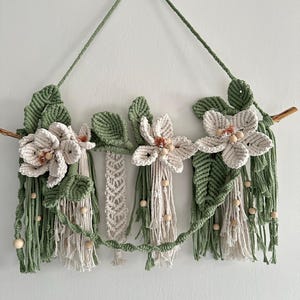 Op de afbeelding: Een macramé wandhanger met drie crèmekleurige bloemen met bruine centra en groene bladeren. De bloemen zijn bevestigd aan een houten tak met groene macramé accenten en hangende franjes. Houten kralen zijn verwerkt.