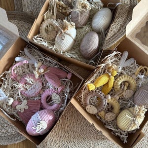 Op de afbeelding: Drie open kartonnen dozen gevuld met paasdecoraties. Elke doos bevat decoratieve eieren, konijntjes en andere ornamenten in tinten wit, roze, geel en bruin, genesteld in versnipperd papier.