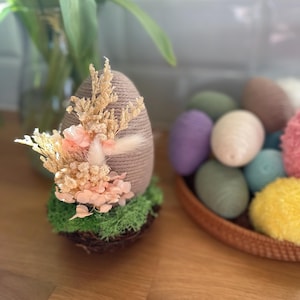 Op de afbeelding: Een decoratief paasei omwikkeld met touw, versierd met gedroogde bloemen en mos, staat op een houten oppervlak. Een mand met kleurrijke, met garen omwikkelde eieren op de achtergrond suggereert een lente- of Paasthema.