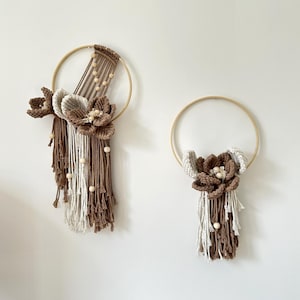 Op de afbeelding: Twee macramé wandkleden. Elk heeft een houten hoepel met een gehaakt bloemdesign in bruin- en crèmetinten. De bloemen hebben houten kralen accenten en franjes.