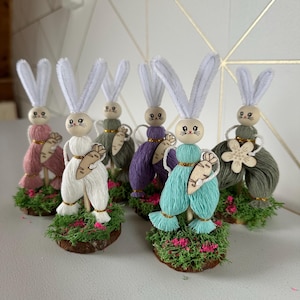Peut inclure: Plusieurs figurines de lapins de Pâques faits à la main. Chaque lapin a une tête en bois, un corps en fil et des oreilles en cure-pipe. Ils tiennent des carottes en bois et sont posés sur une base de mousse et de fleurs roses.