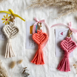 May include: Three macrame heart keychains in cream, coral, and pink, each featuring a gold-tone lobster clasp, a matching satin ribbon, and a decorative card. The cream heart has a yellow ribbon, the coral heart has a pink ribbon, and the pink heart has a pink ribbon.