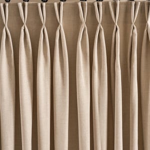 Può includere: Tende in lino beige con un design a pieghe, appese a un'asta di metallo nero con anelli circolari. Le tende sono ben piegate, creando linee verticali. Il tessuto ha un aspetto strutturato, aggiungendo profondità.