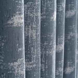 Puede incluir: Primer plano de cortinas azules texturizadas con detalles blancos. La tela tiene una apariencia desgastada y moteada, creando una textura visual. Los pliegues verticales de las cortinas son visibles, agregando profundidad y dimensión al diseño. Las cortinas parecen estar hechas de una tela pesada.