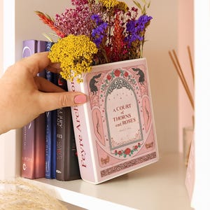 Könnte beinhalten: Eine rosa buchförmige Vase mit dem Text "A Court of Thorns and Roses" und floralen Illustrationen, gefüllt mit bunten Trockenblumen. Die Vase wird in der Nähe eines Regals mit anderen Büchern gehalten.
