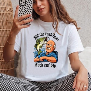 May include: White t-shirt featuring a graphic of a man and a fish with the text "Hit the road Dude Lock em' Up". The illustration is in shades of blue, orange, and green.