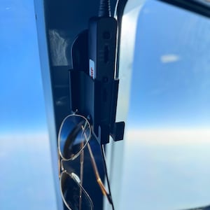 May include: Aviator sunglasses with gold frames and dark lenses hanging from a black holder. A black device with the word "MOP" is mounted on the dashboard. The background shows a clear blue sky.