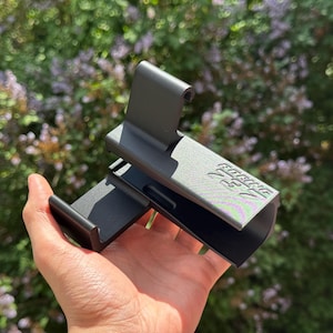 May include: A matte black plastic phone mount featuring a hook-shaped top and a wide base, with the text "BOEING 737" embossed on the side. The device has a textured surface and a rigid, angular construction designed to hold a mobile phone securely.