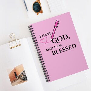 May include: A pink spiral-bound notebook with the text "I HAVE GOD, AND I AM BLESSED" in black and pink. A pink pen illustration is next to the word "GOD". The notebook is open on a white surface, with a gold paper clip and sunglasses nearby.