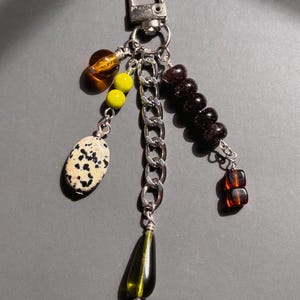 May include: A silver-toned metal keychain featuring a central chain link and multiple dangling charms. The charms include a spotted dalmatian-print stone, two bright yellow beads, a dark amber bead, a row of dark brown beads, and a dark green teardrop.