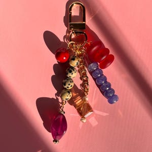 May include: A gold-tone keychain with various colorful beads and charms. The charms include a red apple-shaped bead, a string of dalmatian-patterned beads, a clear orange crystal, a purple teardrop bead, and a red, stacked disc charm.