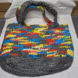 May include: A handmade tote bag with a woven design. The bag has a grey base and handles, with a colourful pattern of red, yellow, blue, and white stripes across the body. The bag is made from recycled materials.
