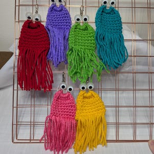 May include: A collection of handmade, colorful crochet earrings. Each earring features a textured, fringed body in red, purple, green, teal, pink, and yellow, topped with googly eyes. The earrings are displayed on a copper-colored grid.