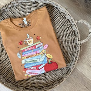May include: A tan t-shirt featuring a graphic of a stack of books with titles including Dragonfly Inn, Weston Bakery, Stars Hollow Books, and Doose's Market. The design includes a coffee mug labeled Luke's, a pumpkin, donuts, and autumn leaves.