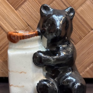 Puede incluir: Una figura de cerámica de un oso negro abrazando un recipiente rectangular blanco con un borde marrón. El oso es brillante y detallado, con sus patas alrededor del recipiente. El recipiente tiene una apariencia ligeramente envejecida.