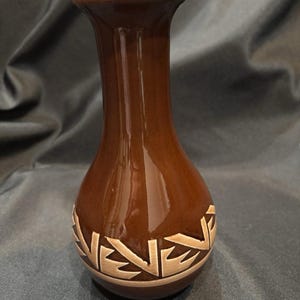 May include: A brown ceramic vase with a glossy finish. The vase features a decorative band of beige geometric shapes near the base. The vase has a slender neck and a flared rim, suitable for displaying small flowers or decorative stems.