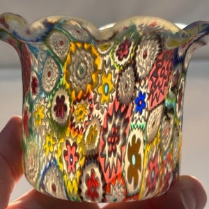 Puede incluir: Una taza de cerámica blanca con asa negra y el texto "But First, Coffee" en negrita negra. La taza se encuentra sobre un fondo borroso, sugiriendo un ambiente acogedor.