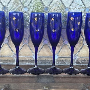 Op de afbeelding: Zes kobaltblauwe champagneflutes met gouden sterrenpatronen. De glazen hebben lange stelen en afgeronde kelken. De flutes zijn in een rij gerangschikt en tonen hun elegante ontwerp. De achtergrond is een glas-in-loodraam.