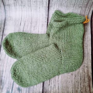 Op de afbeelding: Een paar saliegroene gebreide sokken. De sokken zijn gemaakt van een getextureerd garen en hebben een geribbeld ontwerp op de been en voet. De bovenkant van één sok heeft een omgeslagen manchet met een klein oranje garendetail.