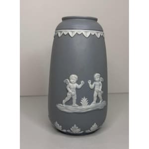 May include: A gray vase with white relief detailing. The vase features a decorative band at the top and bottom, and two cherub figures in the center. The vase is approximately 8 inches tall.