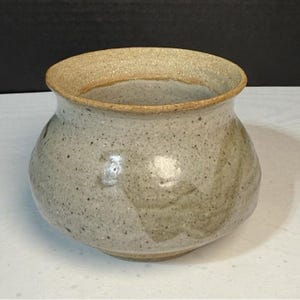May include: A speckled, handmade ceramic pot with a wide opening and rounded body. The pot is primarily a light gray color with darker speckles and a tan rim. The overall shape is a squat, rounded form.