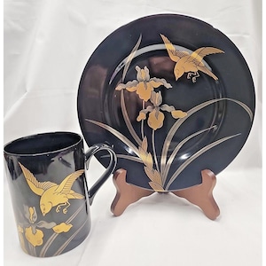 May include: A black ceramic mug and plate set with gold bird and floral designs. The plate is propped on a wooden stand. The mug has a curved handle. The plate and mug have a matching design of gold birds and irises.